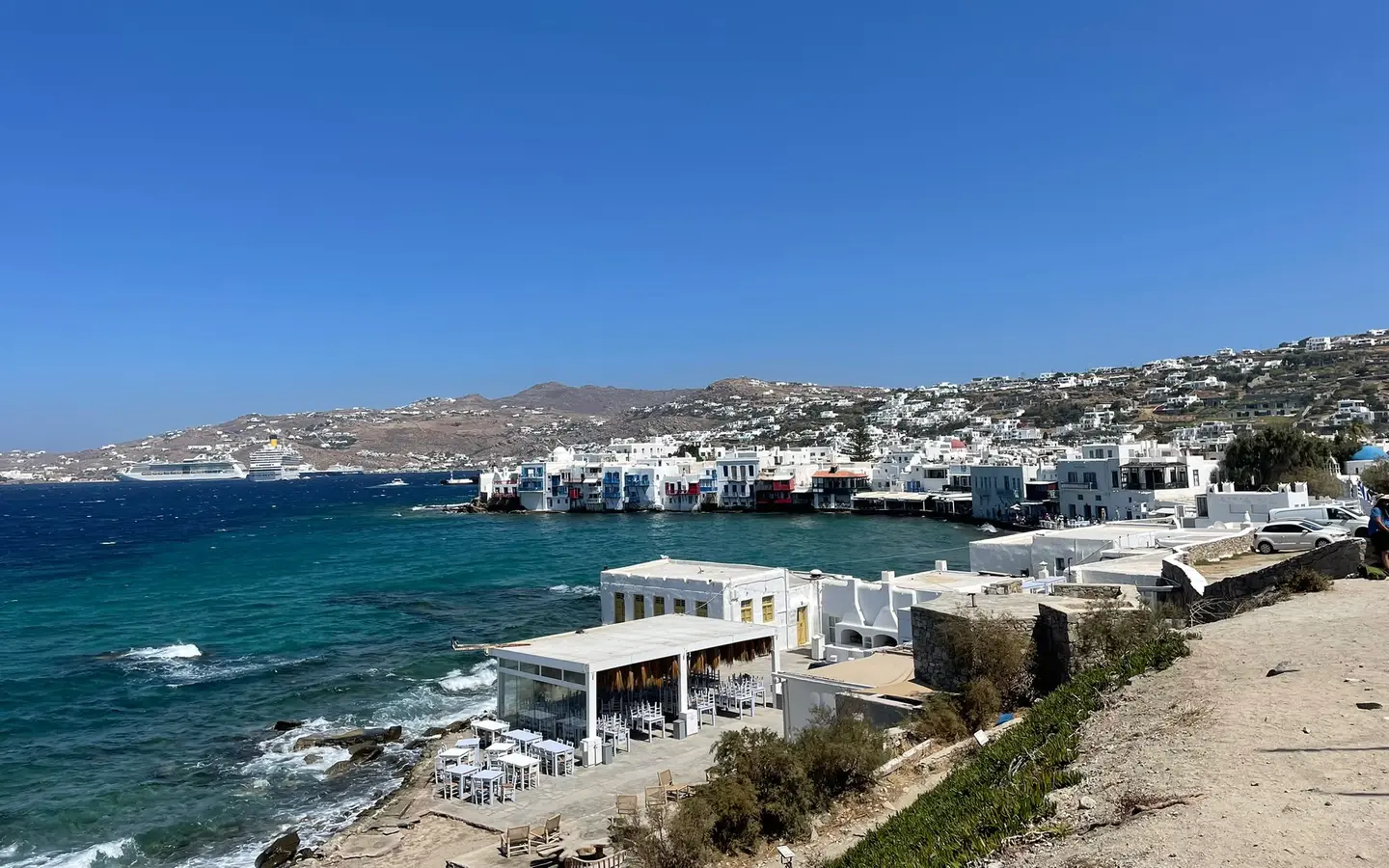 Little Venice auf Mykonos mit weißen Häusern am Wasser, im Hintergrund liegen Kreuzfahrtschiffe auf Reede.