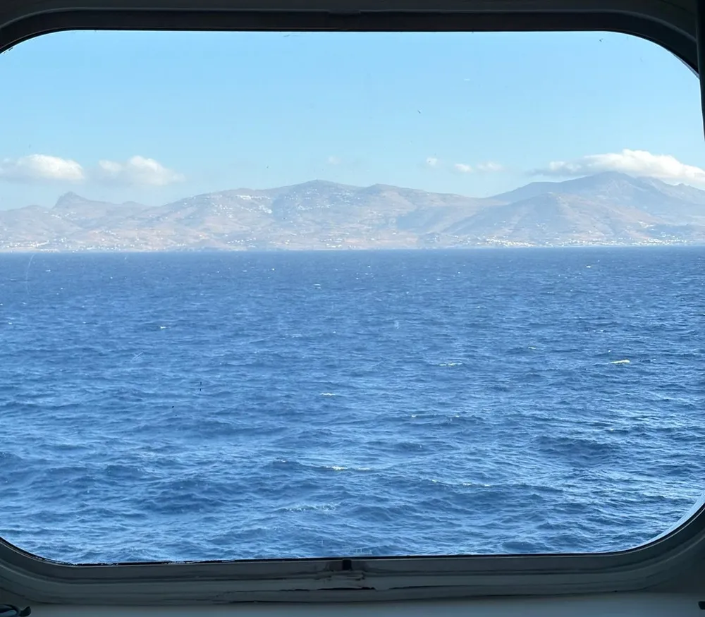 La vista dalla finestra di una cabina esterna: mare blu e un'isola all'orizzonte, con il letto in basso nell'inquadratura.
