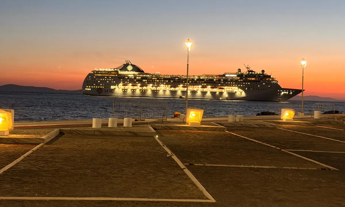 MSC Lirica illuminata all'ancora davanti a Mykonos dopo il tramonto, con una piazza del porto vuota e lampioni in primo piano.