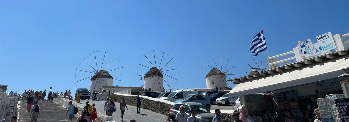 Die Windmühlen von Kato Mili auf Mykonos über einer belebten Straße, rechts eine griechische Flagge.