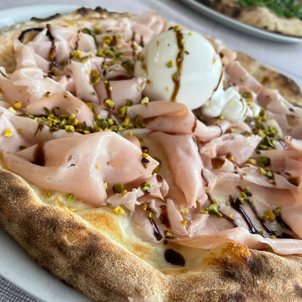 Pizza mit Mortadella, Burrata, Pistazien und Balsamico auf einem Teller, im Hintergrund eine zweite Pizza.