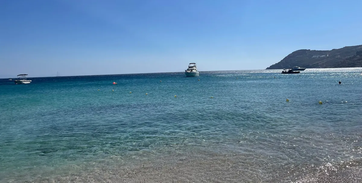 Klares, türkisfarbenes Wasser an einem Strand auf Mykonos, draußen liegen ein paar Boote vor Anker.